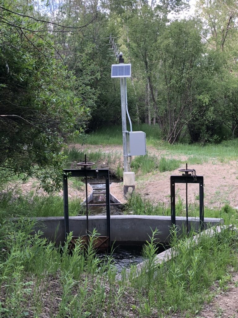 Augmentation station with solar panel, satellite telemetry antenna, and equipment enclosure standing above a measured headgate structure