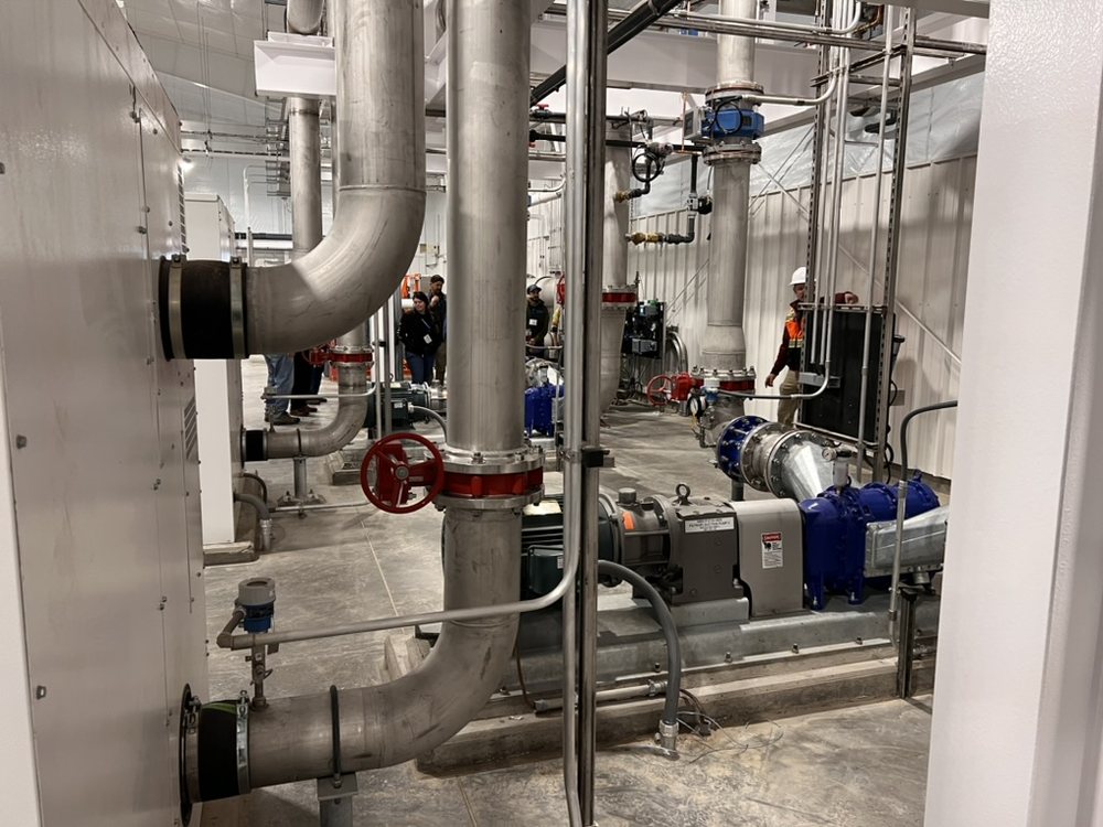 Stainless-steel process piping, gate valves, and pumps on the floor of a water treatment plant, with crew in hard hats and hi-vis vests